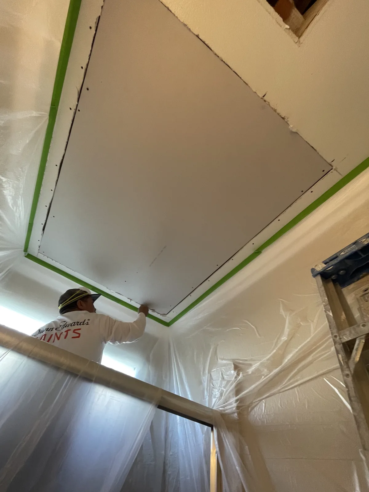Rectangular ceiling drywall patch bordered by green masking tape above plastic-covered walls